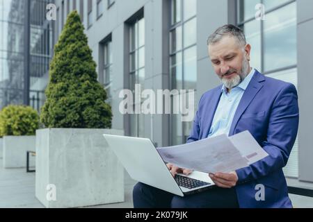 Senior handsome successful male engineer architect in suit works with laptop, documents, plan, drawings. Sitting on a bench near the office center, holding papers, looking around, smiling. Stock Photo