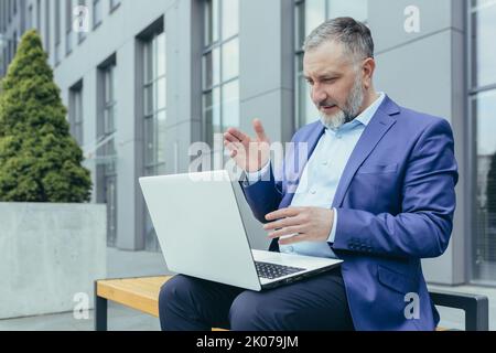 Successful senior male businessman conducts a video conference from a laptop. Sitting outside in a suit on a bench near an office center, waves his hands, explains. Stock Photo