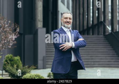 Portrait of a successful senior male businessman in a business suit standing confidently near the office center in full height, arms crossed, smiling. Stock Photo