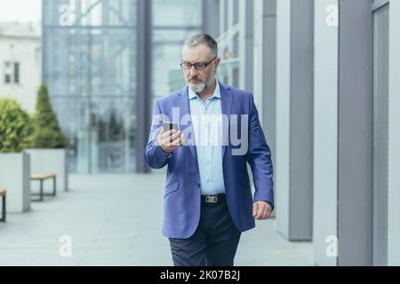 Serious gray-haired senior handsome man businessman in suit and glasses walking near office center on street, holding mobile phone, typing message, checking mail. Stock Photo