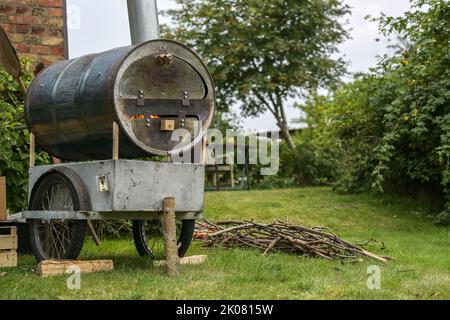 Homemade mobile pizza oven from an old metal barrel on a recycled chassis in a garden, fire in the combustion chamber and firewood on the lawn, copy s Stock Photo