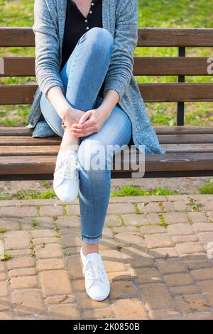 White sneakers on female legs in blue jeans on a blue bench and green plant background. Stock Photo