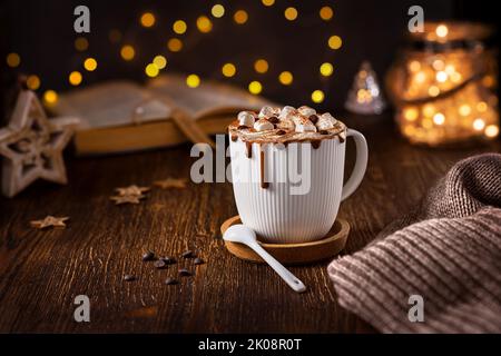 Hot chocolate with marshmallows in a white cup on a wooden table with a book and a garland in the background. Stock Photo