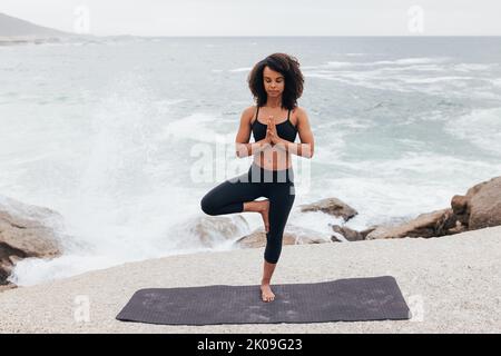Woman with curly hair practicing Tree Pose by seaside. Young female with closed eyes doing Vrksasana at evening. Stock Photo