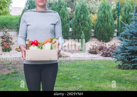 Young woman holding in hands wooden box full of fresh harvested vegetables. Organic tomatoes, pattypan, carrot, peppers, parsley, peas. Copy space. Stock Photo