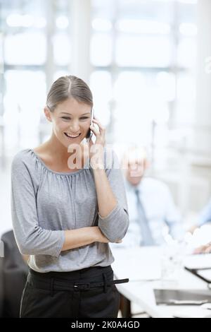 Staying in touch with her clients. A beautiful woman using her cellphone in the office with her colleagues in the background. Stock Photo