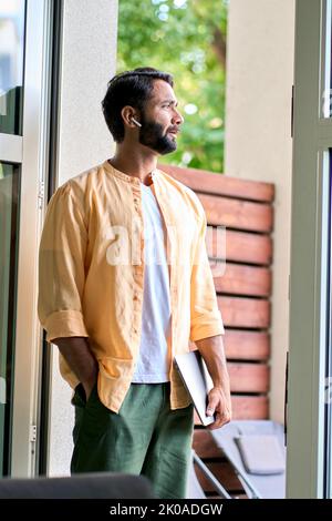 Thoughtful indian man holding laptop standing at doorway and thinking. Stock Photo