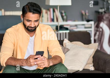 Indian man sitting at home on couch using smartphone watching videos. Stock Photo