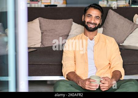 Happy indian man looking at camera drinking coffee sitting on sofa at home. Stock Photo
