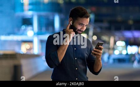 Indian man wearing earbud holding cell phone at night using smartphone. Stock Photo