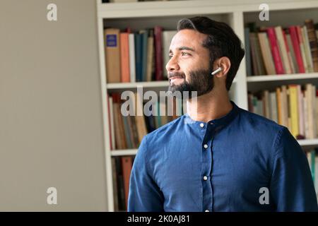 Thoughtful indian man wearing earbuds listening podcast looking through window. Stock Photo