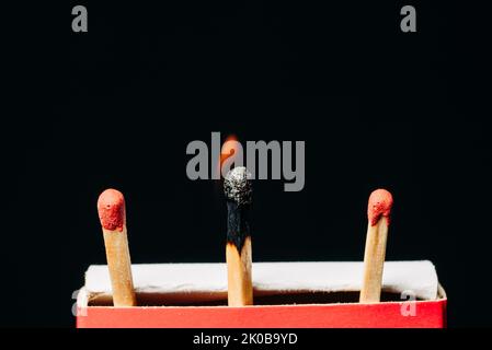 Lit match producing a small flame next to two unlit matches against a black background. Stock Photo