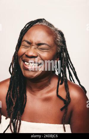 Joyful mature woman laughing cheerfully while wrapped in a bath towel. Refreshed woman with dreadlocks standing against a white background. Happy midd Stock Photo