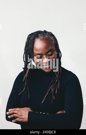 Professional woman with dreadlocks smiling happily while wearing eyeglasses. Cheerful mature woman standing against a grey background with her arms cr Stock Photo