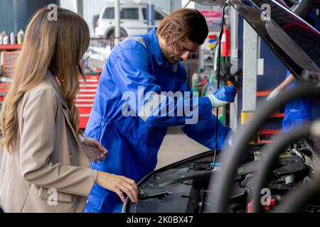 Young man mechanic talking with customer for examining trouble and repairing engine while using oil dipstick checking oil level in engine of car in ga Stock Photo