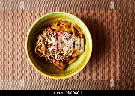 Udon stir-fry noodles with chicken and sesame seeds in green bowl on noisy brown background, on table mat. Top view Stock Photo