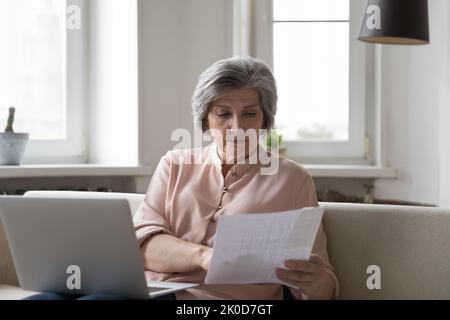 Focused senior elder accountant woman checking paper invoice at laptop Stock Photo