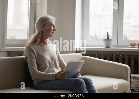 Positive thoughtful senior 70s lady holding tablet computer Stock Photo