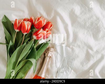 Bunch of red tulips, rose wine and champagne glasses on white bed sheets. Valentines day, date, march 8 concept. Spring flowers and bottle aesthetics. Top view or flat lay. Copy space Stock Photo