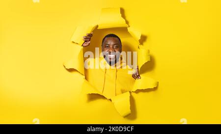 Portrait of happy black man standing in torn paper hole, looking and smiling at camera through yellow background Stock Photo