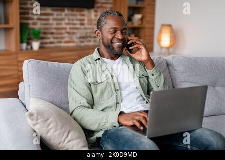 Smiling busy middle aged african american businessman in casual calling by phone and typing on laptop Stock Photo