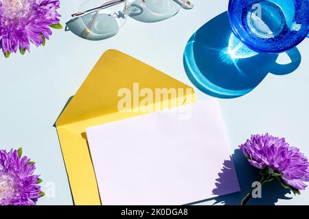 White blank card, yellow envelope, glass of water, eyeglasses and flowers on blue background. Bright stilllife, Top view Flat lay Mockup. Stock Photo