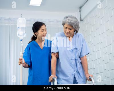 Elderly Asian woman patient trying to walk on walking frame held and carefully supported in arms by caregiver, young polite female assistant nurse in Stock Photo