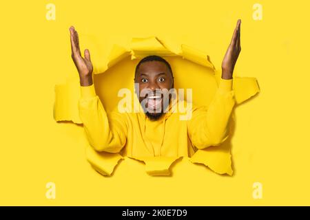 Portrait of excited black man looking through hole in yellow paper, standing with raised arms, screaming happily Stock Photo