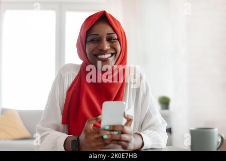 Smiling black lady in hijab sitting at home with cellphone, checking social network, browsing web, video chatting online Stock Photo