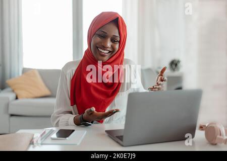 Cheerful black muslim woman in hijab having video chat with friends or online meeting, talking to laptop webcamera Stock Photo