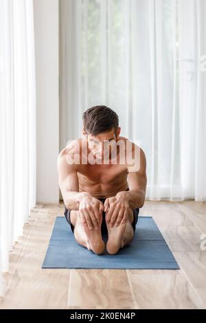Young sporty man doing stretching legs on yoga mat during morning training Stock Photo
