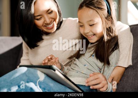 Happy mother using tablet computer while hugging her daughter at home Stock Photo