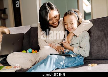 Happy mother using tablet computer while hugging her daughter at home Stock Photo