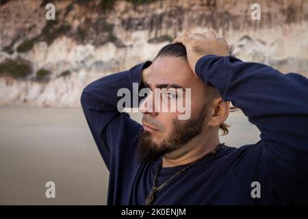Middle-aged white man with a beard and his hands on his head looking at the camera from the side on the beach. Stock Photo