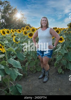 Woman with light brown hair and white shirt standing in the middle of a yellow, green, and brown sunflower field located outside of Lawrence Kansas Stock Photo
