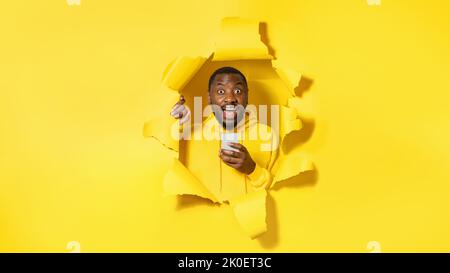 Portrait of surprised black man using cellphone, reading shocking news, standing in torn yellow paper hole Stock Photo