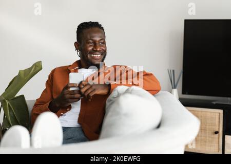 Excited black guy chatting on cellphone or surfing internet, sitting on sofa at home, looking aside at copy space Stock Photo