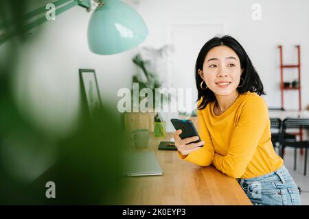 Smiling young asian woman holding mobile phone while working on laptop at home Stock Photo