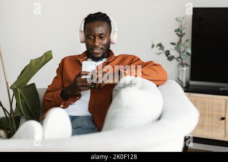 Relaxed black man watching movies on smartphone, wearing headset and using cellphone, sitting on sofa, copy space Stock Photo