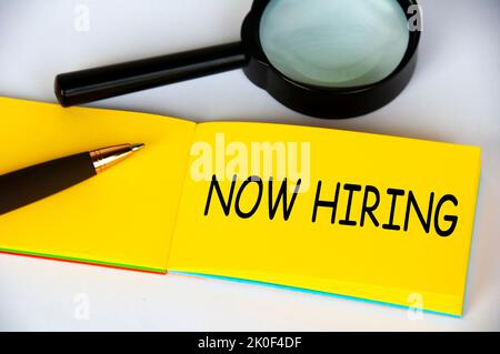 Now hiring text on yellow notepad with magnifying glass and pen on white background. Employment concept. Stock Photo