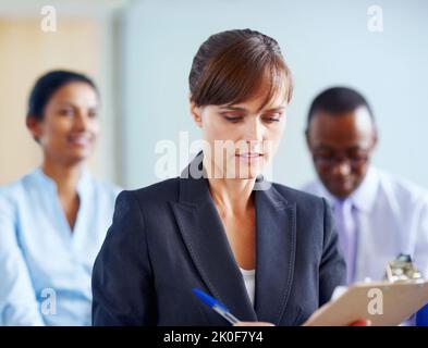Female executive taking down notes. View of female executive making notes with colleagues in background. Stock Photo