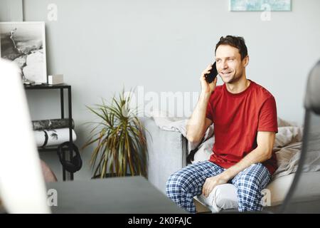 Young man having conversation on mobile phone while sitting on bed in his bedroom Stock Photo