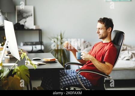 Young man eating sandwich and drinking coffee while sitting on chair in front of computer and watching video Stock Photo