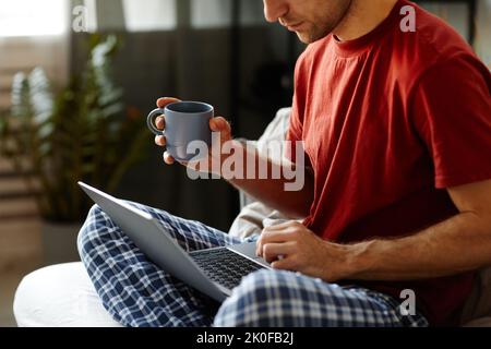 Close-up of man drinking coffee and typing on his laptop while sitting on bed at home Stock Photo