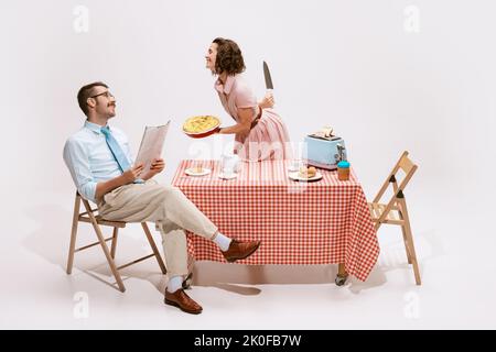 Portrait of couple sitting at the table, having breakfast isolated on white background. Movie remake. Wife hiding knife behind back Stock Photo