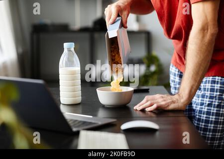 Close-up of young man putting cornflakes in bowl to make them with milk for breakfast at table with laptop Stock Photo