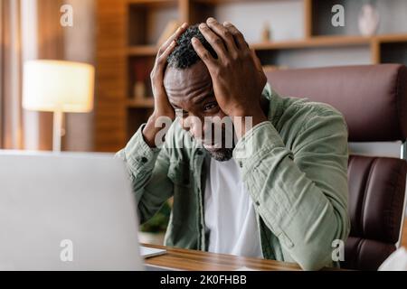 Sad frustrated adult african american guy in casual hold his head with hand watch at laptop, suffer from stress Stock Photo