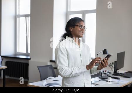 Happy thoughtful Indian employee man holding smartphone, standing at workplace Stock Photo