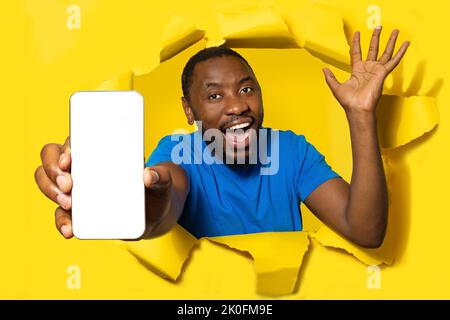 Overjoyed black man with open mouth showing smartphone with blank screen through torn hole in yellow paper, mockup Stock Photo