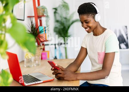 Happy young african woman with laptop using mobile phone while sitting at desk Stock Photo